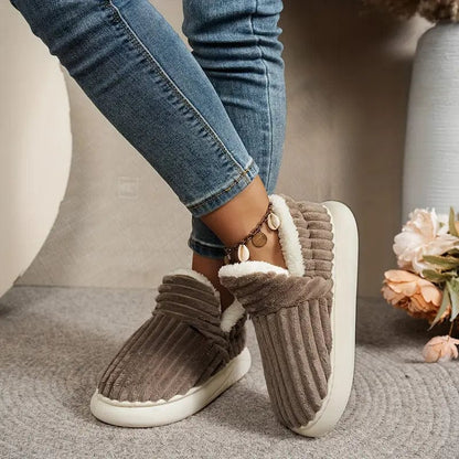 Brown ribbed slippers worn by a model in jeans with legs crossed, beside a vase of flowers. 9