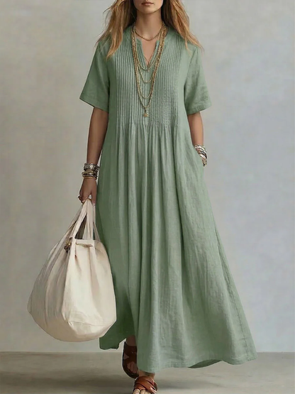 Model wearing the short-sleeved V-neck dress in soft sage green, with pintucked bodice, layered gold necklaces and a cream tote bag. 2