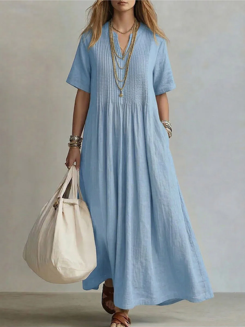 Model wearing the short-sleeved V-neck dress in light sky blue, with pintucked bodice, layered gold necklaces and a cream tote bag. 3