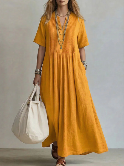 Model wearing the short-sleeved V-neck dress in warm mustard yellow, with pintucked bodice, layered gold necklaces and a cream tote bag. 4