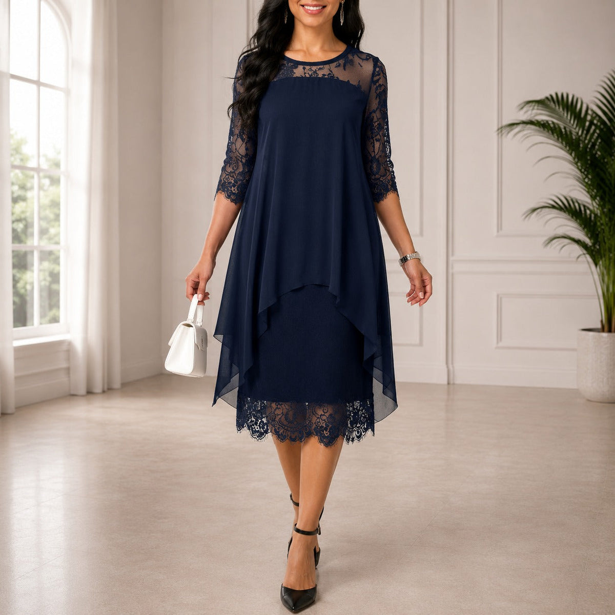 Woman in a navy layered chiffon dress with lace sleeves and scalloped lace hem, holding a white handbag in a bright room. 1