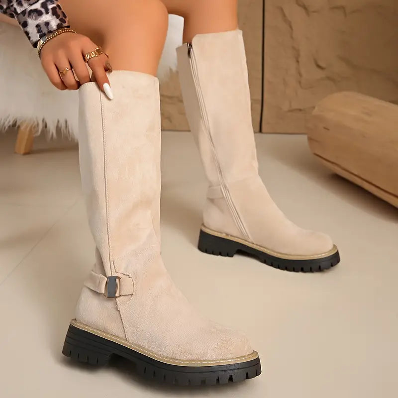 Model wearing cream tall boots with a buckle strap at the ankle and a side zip on the other boot. 9