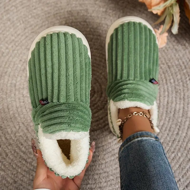 Green ribbed slippers seen from above, one held in a hand and one worn, on a woven grey rug. 3