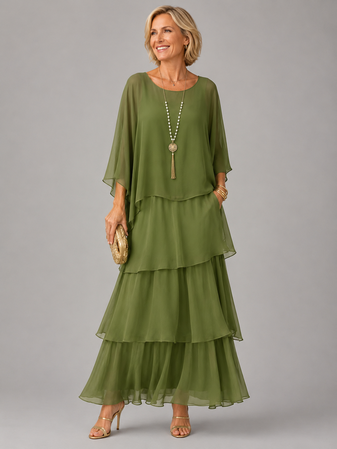 Smiling woman in the olive green tiered chiffon maxi dress with draped cape sleeves, long beaded necklace and gold heeled sandals. 2