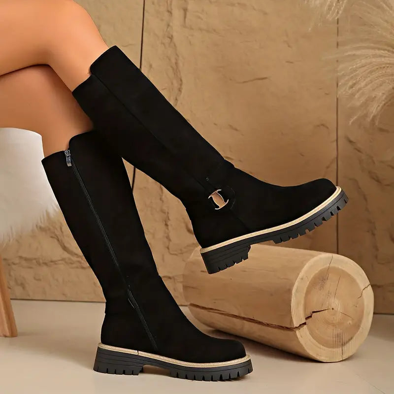 Model with legs crossed in black knee-high boots, showing the inner side zip, small buckle strap and thick treaded sole. 2