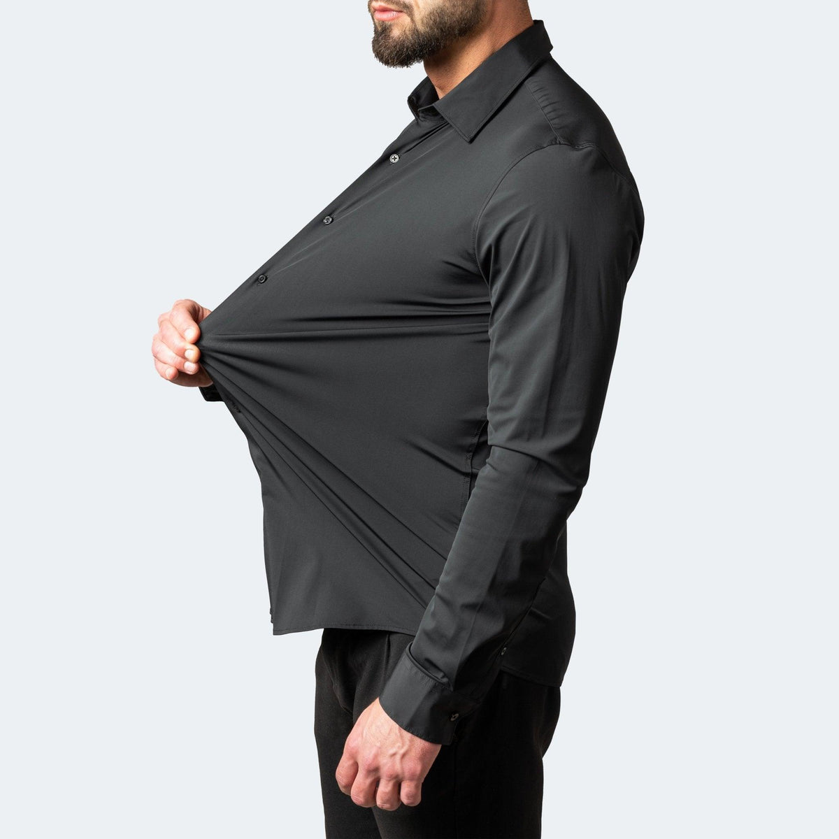 Profile view of a bearded model in a black shirt pulling the front outwards to show how far the fabric stretches. 2