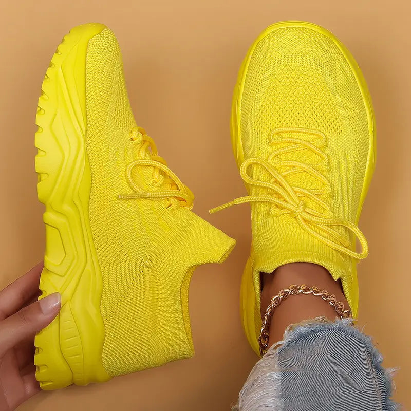 Top-down view of bright yellow knit sneakers with matching chunky yellow soles, one held up and one worn with frayed jeans. 18