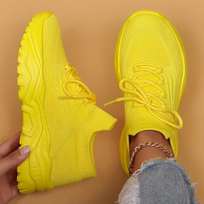 Top-down view of bright yellow knit sneakers with matching chunky yellow soles, one held up and one worn with frayed jeans. 18