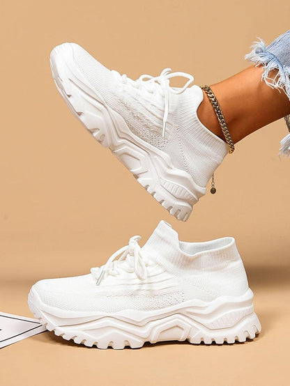 Side view of all-white knit sock-style sneakers with thick sculpted white soles, one raised above the other, gold anklet visible. 7