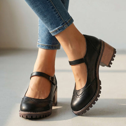Black brogue-detail shoes with buckled ankle strap and chunky stacked heel, worn with rolled blue jeans in a bright indoor setting. 1