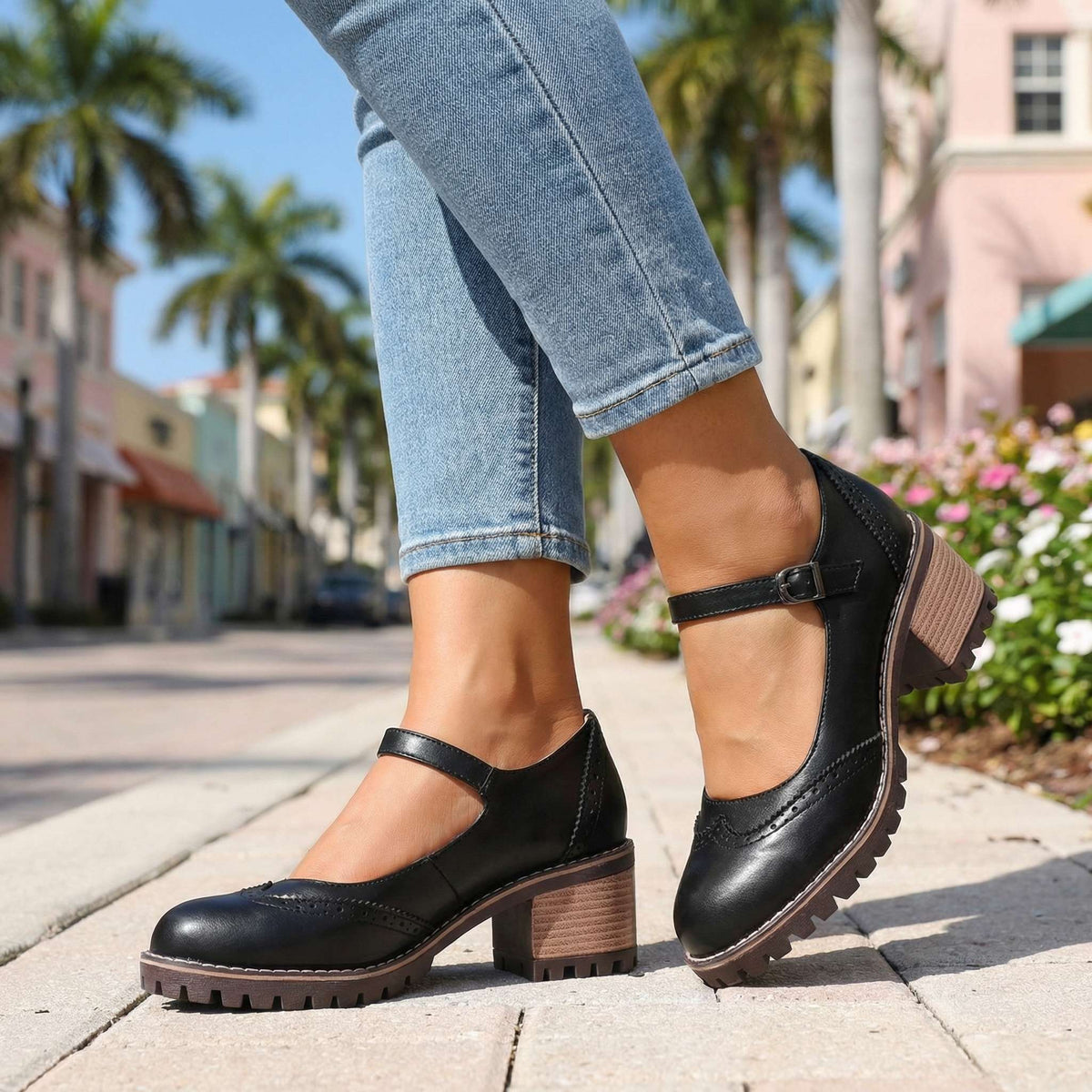 Black brogue Mary Jane heels shown from the side with buckle strap, worn with cropped jeans beside pink flowers outdoors. 7