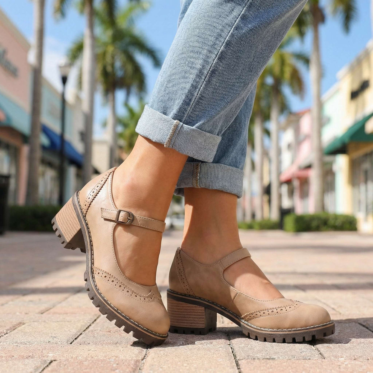Beige pair worn on a palm-lined shopping street, showing the adjustable buckle strap, punched brogue pattern and block heel. 6