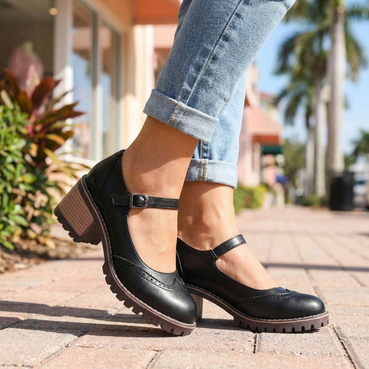 Black Mary Jane block heels with buckled ankle strap, worn with cuffed light jeans on a paved street beside palm trees. 4