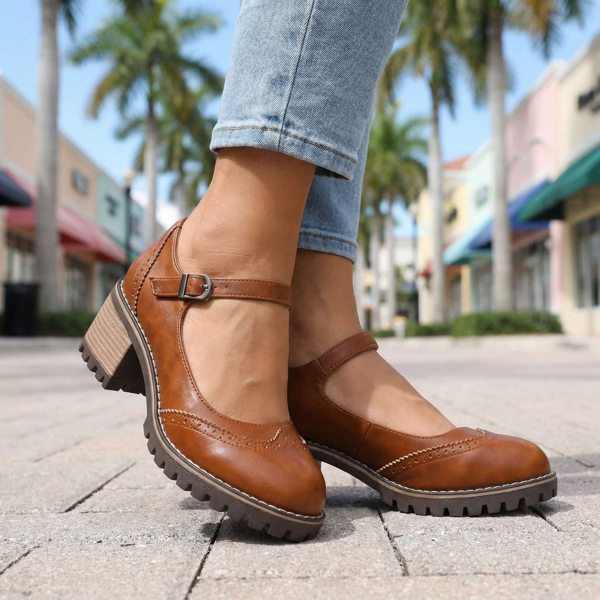Close view of tan brown Mary Jane heels with metal buckle and brogue perforations, worn on a paved outdoor street. 8