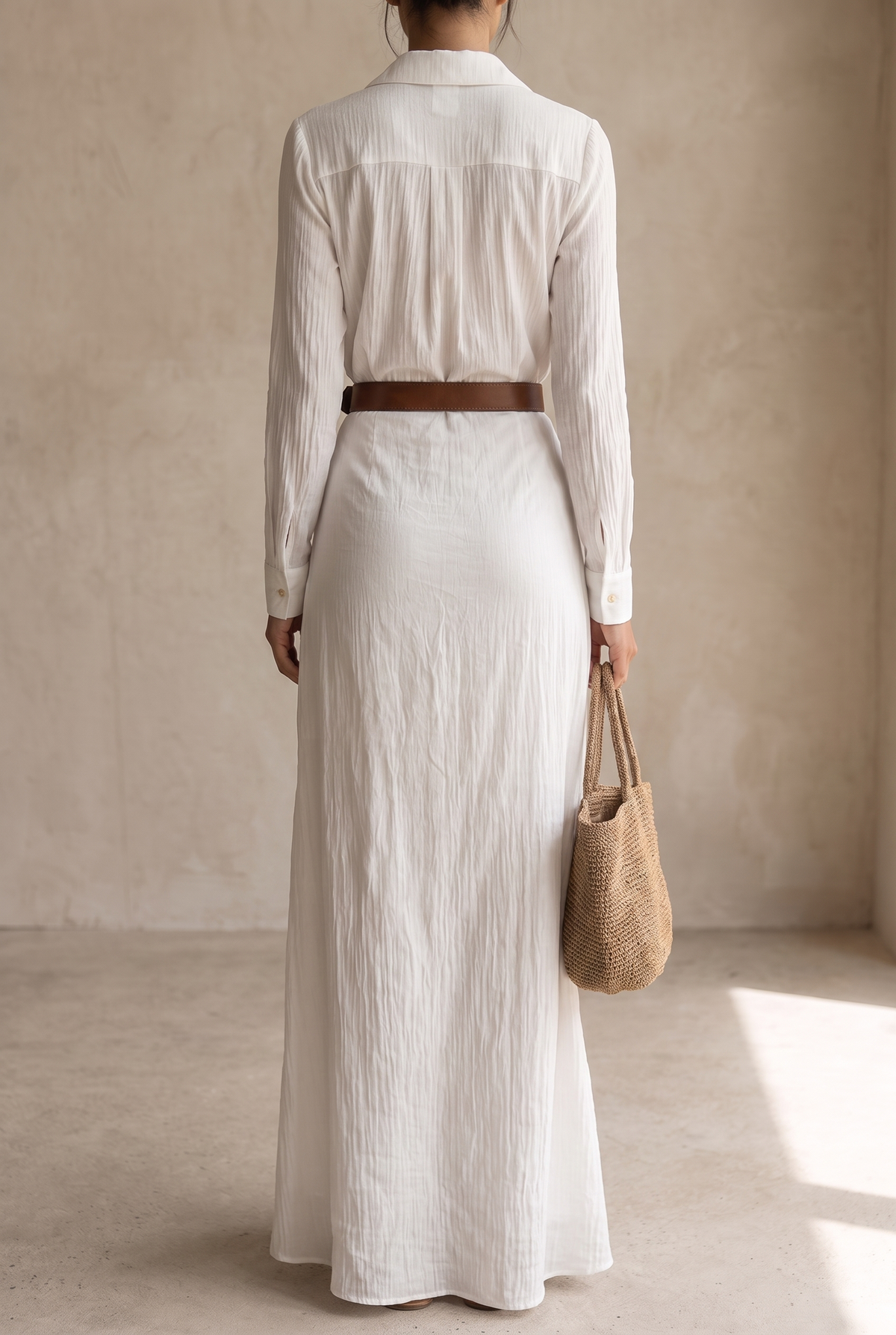 Back view of the white maxi dress, showing the pleated yoke, button cuffs, brown belt and straw tote held in one hand. 11