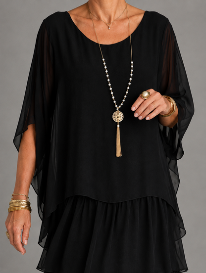 Close-up of the black dress bodice with round neckline and sheer draped sleeves, styled with a pearl and gold tassel necklace. 18