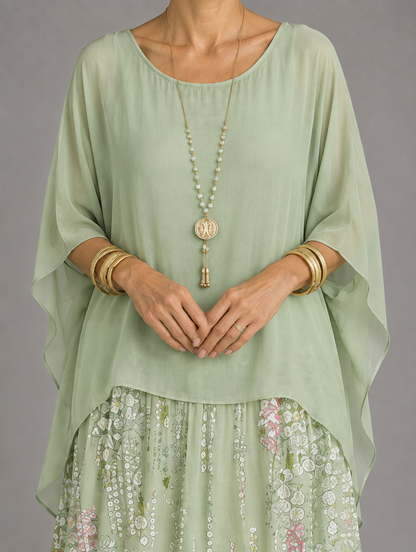 Close-up of a sage green sheer chiffon overlay with wide sleeves, worn with a pendant necklace over a floral tiered skirt. 48