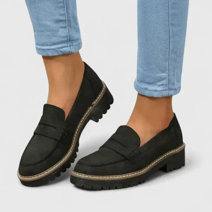 Black suede-look penny loafers with chunky treaded soles and contrast stitching, worn with light blue cropped jeans. 1