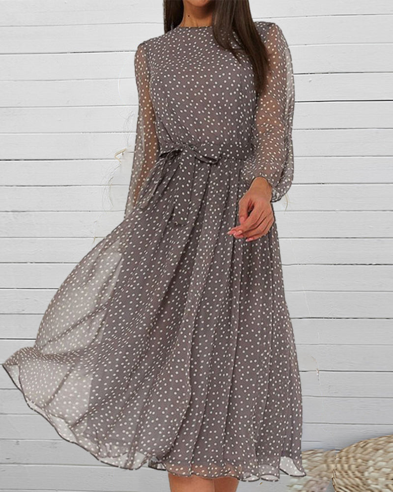 Grey polka dot dress with sheer sleeves and tied waist, worn by a model in front of white wooden panelling. 2