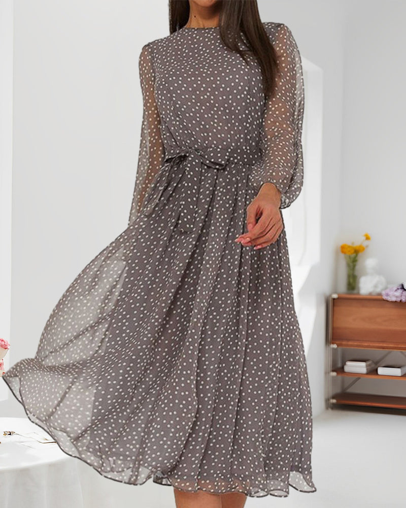 Model in the sheer-sleeved grey polka dot dress, photographed in a bright room with a wooden shelf and yellow flowers. 3