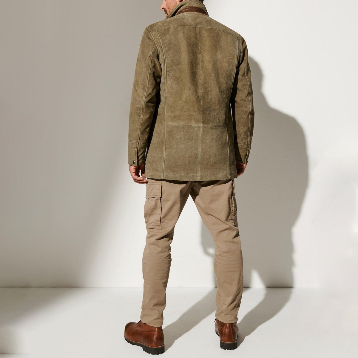 Back view of a man wearing the olive suede jacket with beige cargo trousers and brown leather boots, showing panelled seams. 3