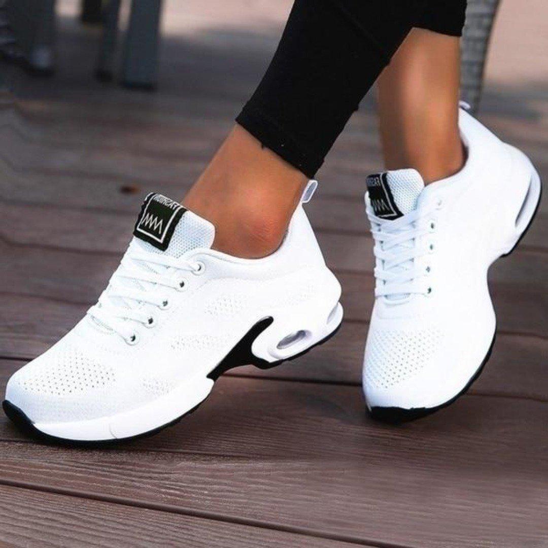 Plain white mesh trainers with a black-edged air-cushion sole and white laces, worn with black leggings on wooden decking. 6