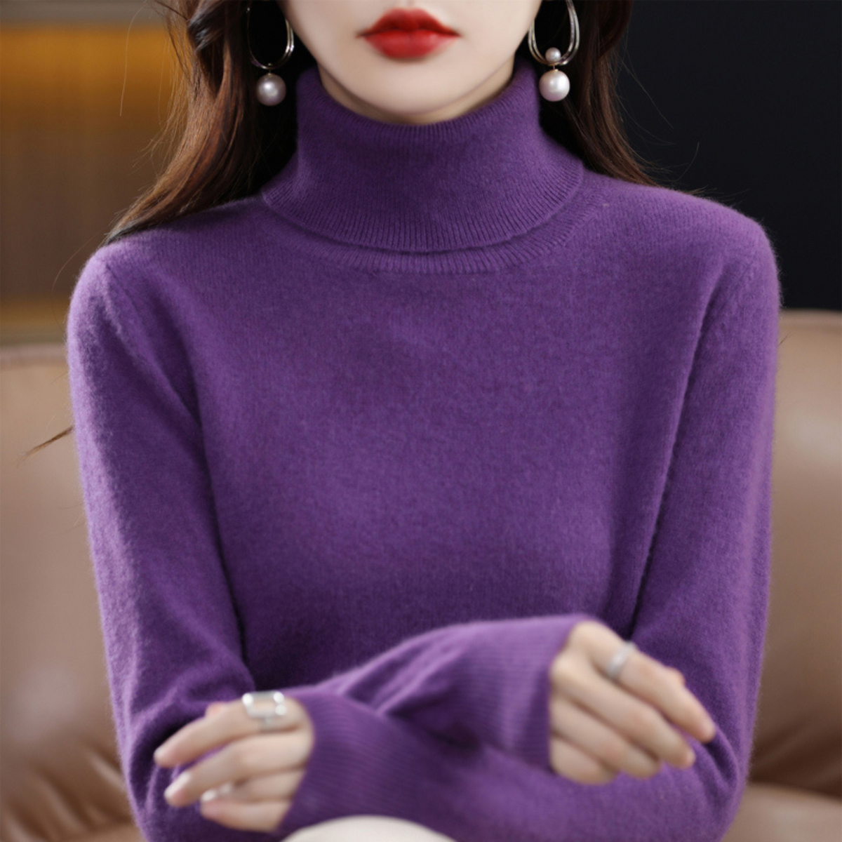 Model wearing the roll neck jumper in deep purple with arms crossed, seated against a tan leather sofa. 5