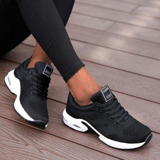 Black mesh lace-up trainers with thick white cushioned soles and visible air units, worn with black leggings on wooden decking. 1