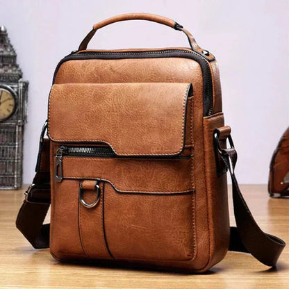 Tan textured shoulder bag with top handle, flap pocket, zip and metal ring, shown on a wooden table beside an ornamental clock. 2