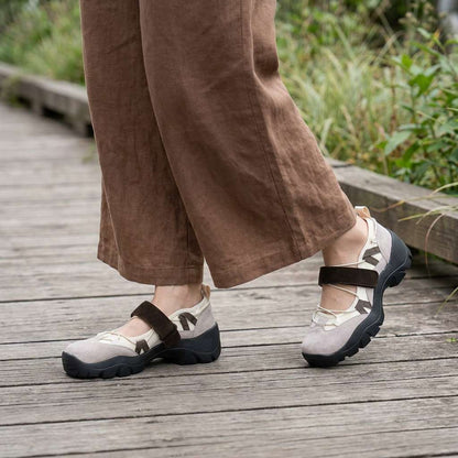 Beige suede shoes with brown straps, worn with wide brown linen trousers on a wooden walkway beside greenery. 11