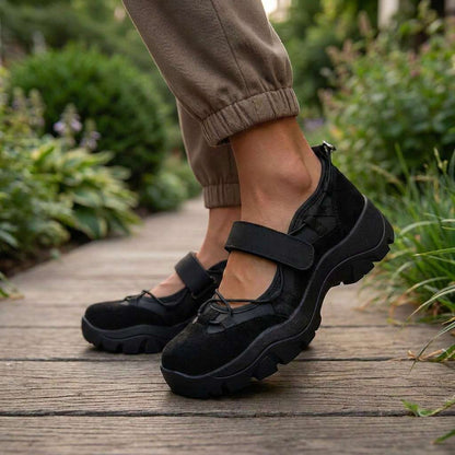 Black suede shoes with a strap worn with khaki cuffed trousers, on a wooden garden path among green plants. 10
