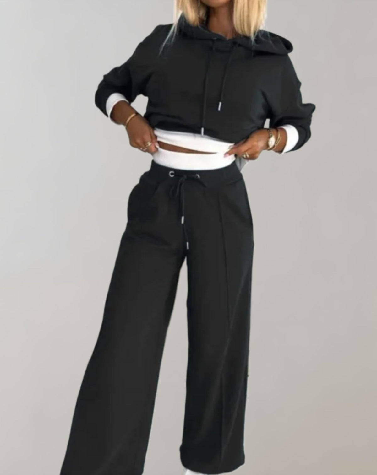 Model in the black cropped hoodie and wide leg trousers, lifting the hem to show the white ribbed waistband layer. 1