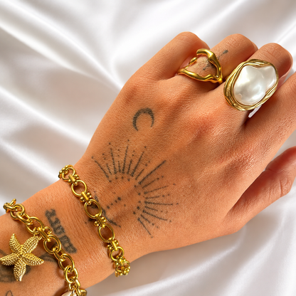 Wavy gold-tone open ring worn on a finger beside a large pearl-style ring and gold chain bracelets, on white satin. 4