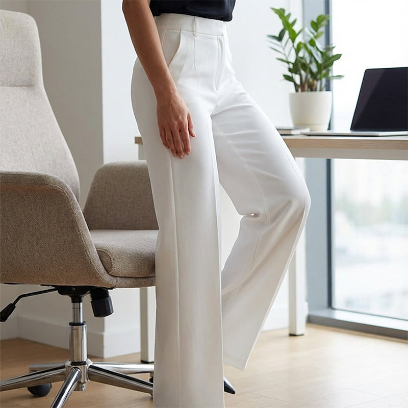 Close crop of a model in white high-waisted trousers with a hand at the pocket, leaning near an office chair and desk. 14
