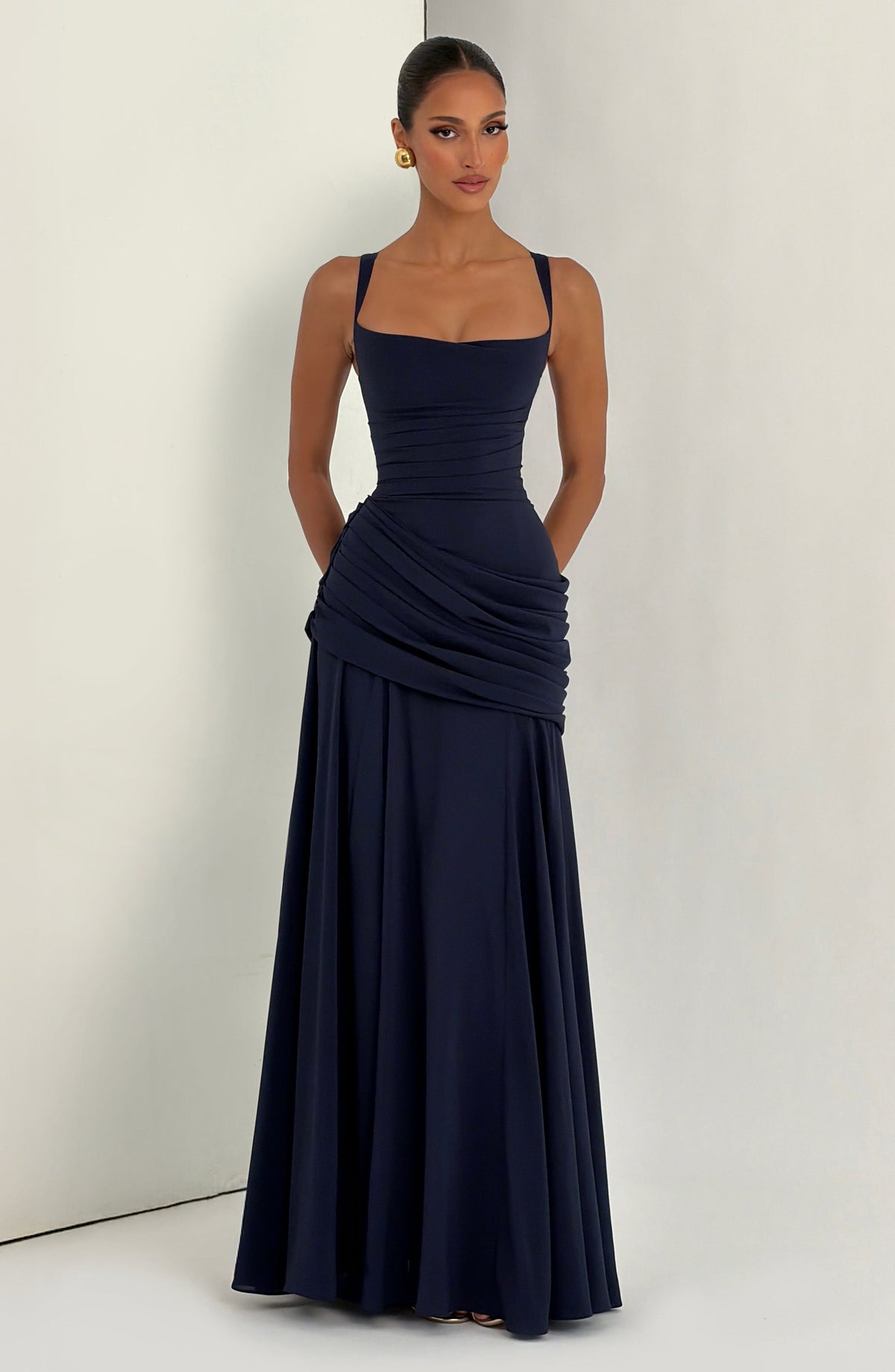 Navy maxi dress worn by a model against a light studio corner, front view with hands behind her back and the skirt flowing. 13