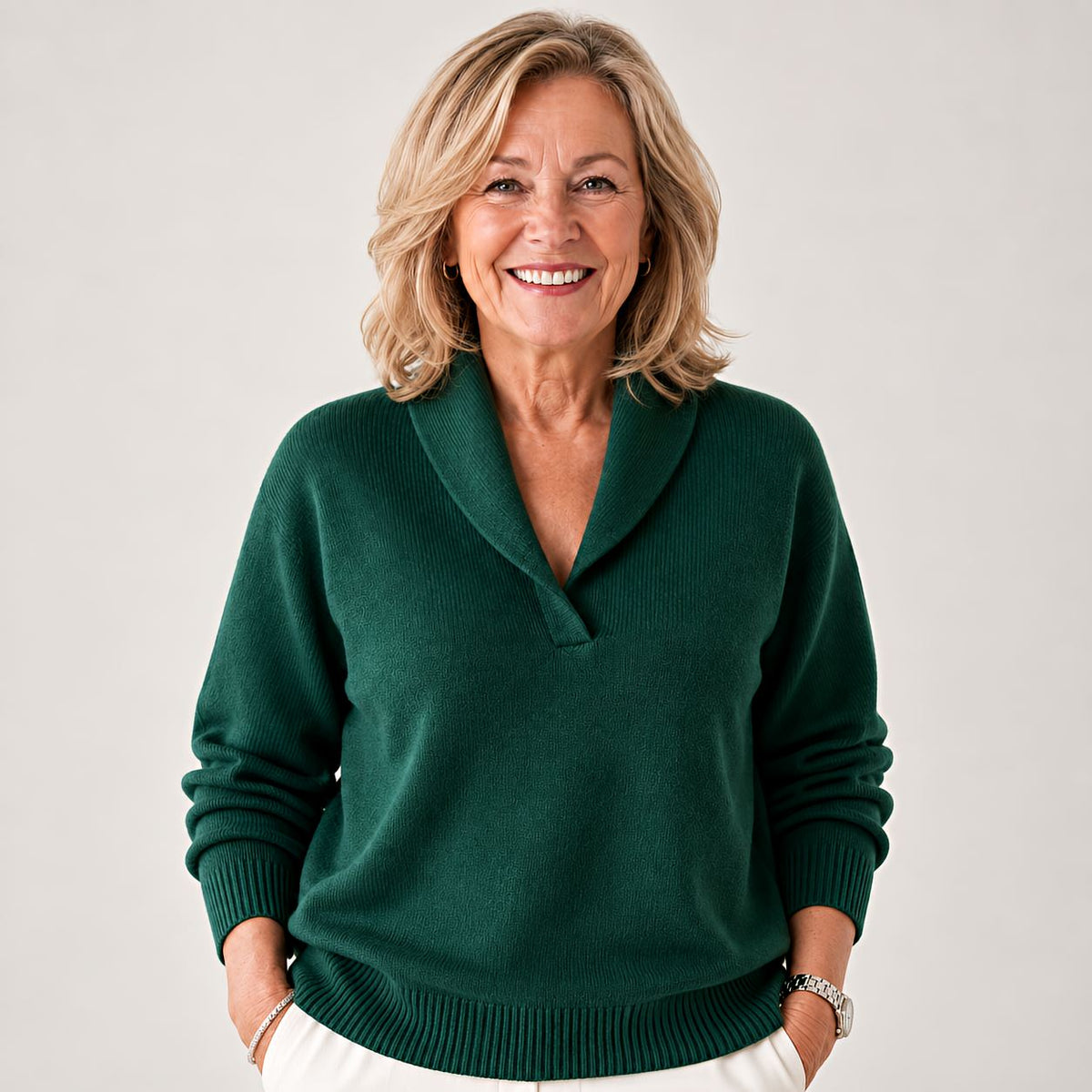 Smiling woman wearing the knitted jumper in dark green, hands in the pockets of white trousers, shown from the front. 1