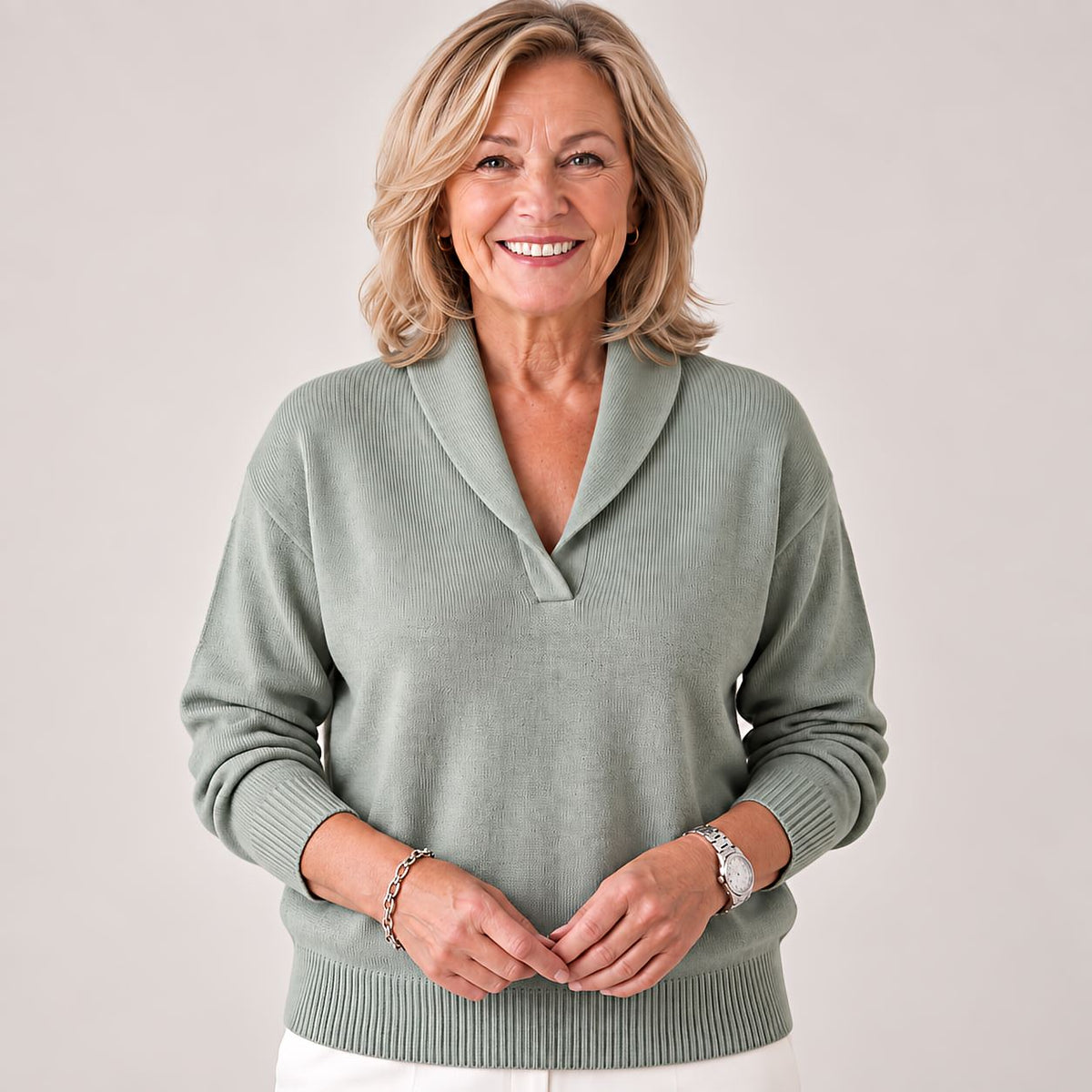 Smiling woman with hands clasped in front, wearing the jumper in sage green with white trousers. 5
