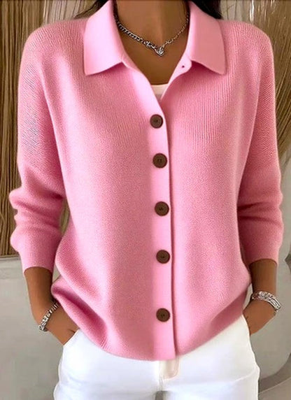 Model wearing the pink version of the knit cardigan, buttoned up with a collar, over a white top and white trousers. 2