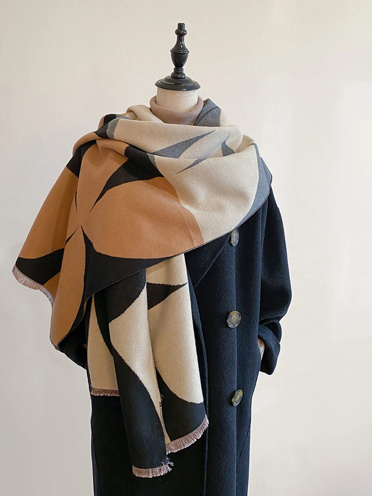 Scarf in camel, cream, black and grey geometric shapes wrapped round a mannequin wearing a long black coat. 2