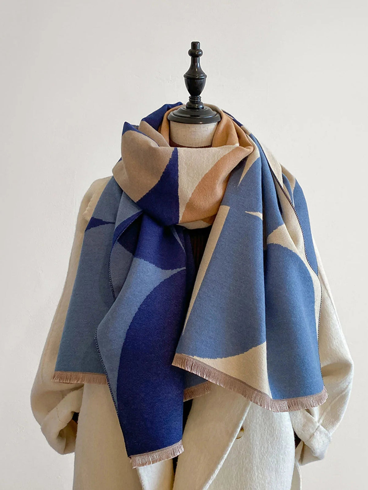 Blue, camel and cream geometric scarf loosely wrapped on a mannequin in a cream coat, fringed ends hanging down. 3