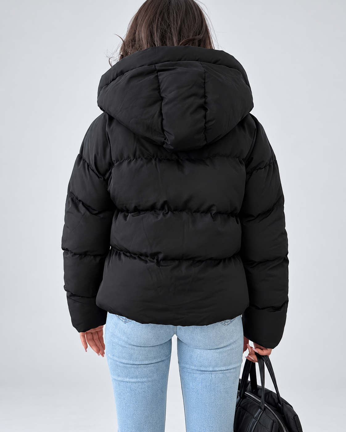 Back view of the black padded jacket on a model, showing the hood and horizontal quilted panels, with light blue jeans and a black bag. 3