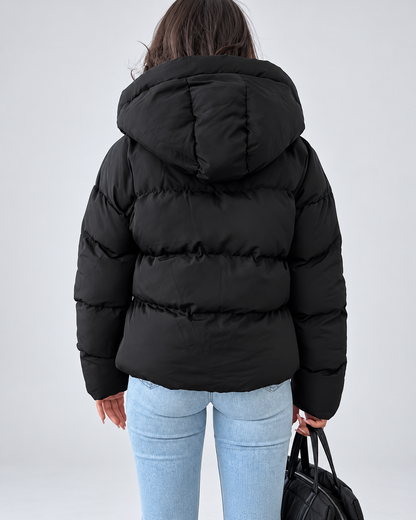 Back view of the black padded jacket on a model, showing the hood and horizontal quilted panels, with light blue jeans and a black bag. 3